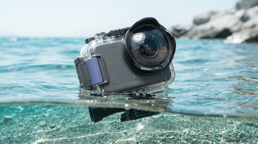 An action camera held half-submerged at the waterline of a clear turquoise sea, droplets beaded on the lens housing, with bright sunlight overhead and a sandy seabed visible below