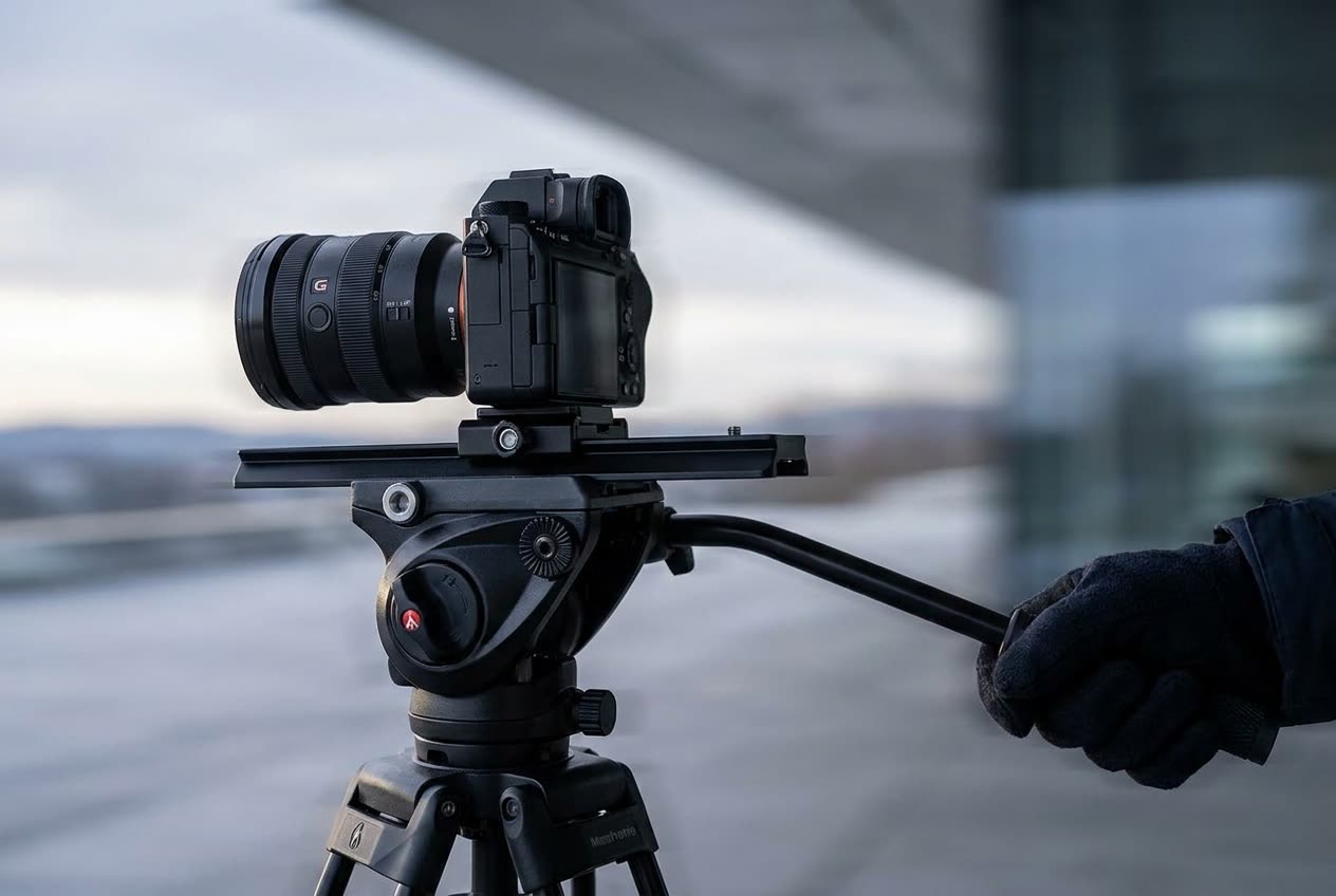 A close view of a fluid video head with a mirrorless camera balanced on its long sliding plate, a pan handle extending to the right, the drag and counterbalance dials labeled on the side of the head