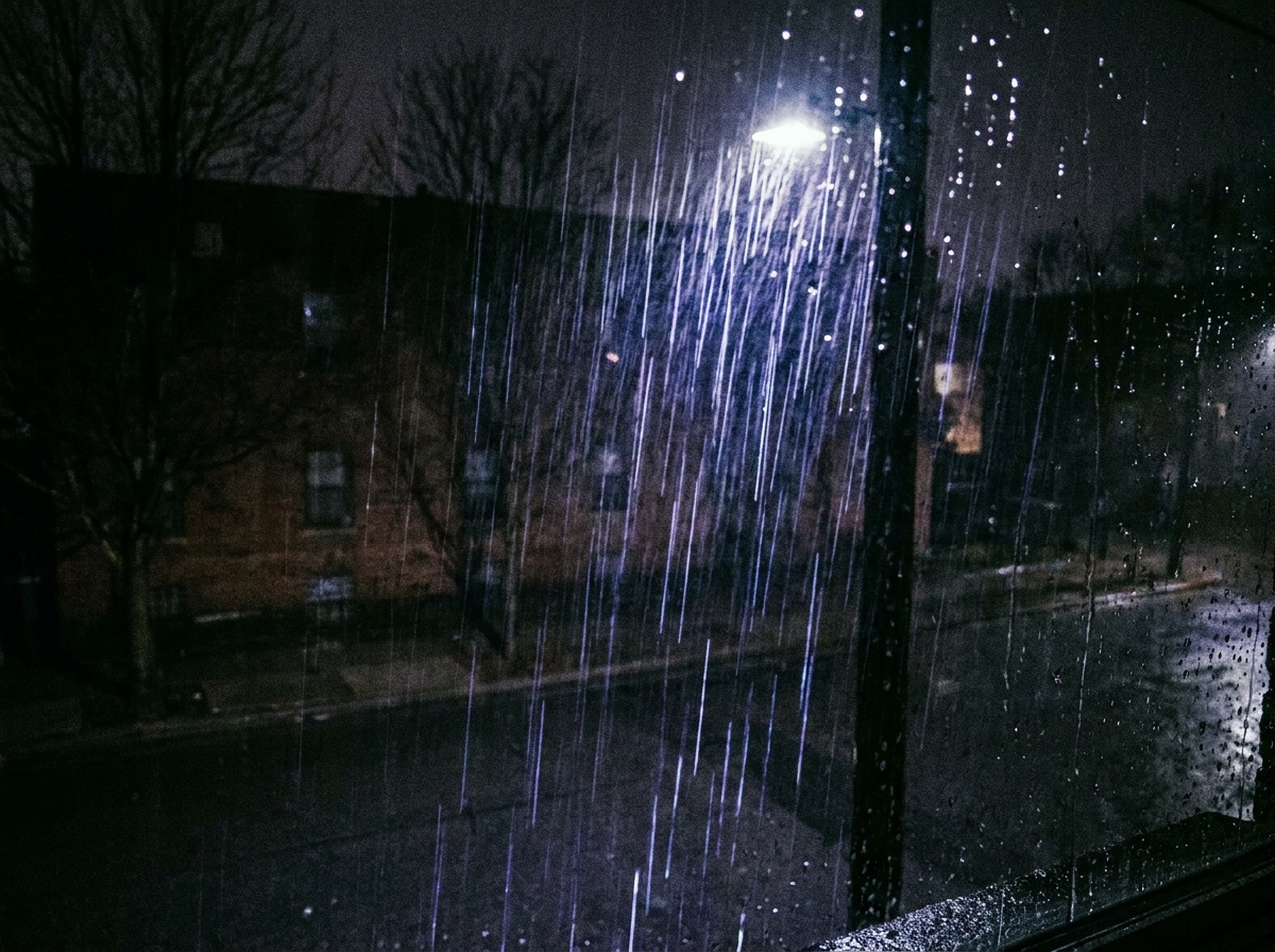 Heavy rain caught in the cone of a single streetlamp at night, the falling drops lit into thousands of bright silver streaks against the deep shadow of the street behind