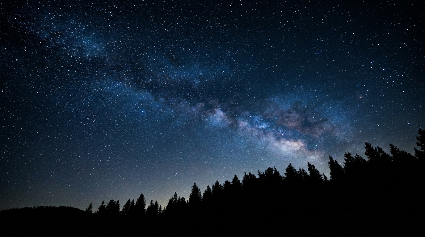 A wide frame of a deep, star-packed night sky glowing with thousands of pinpoint stars over the black silhouette of a pine ridge, the faint band of the Milky Way arcing through and the sky a rich natural blue